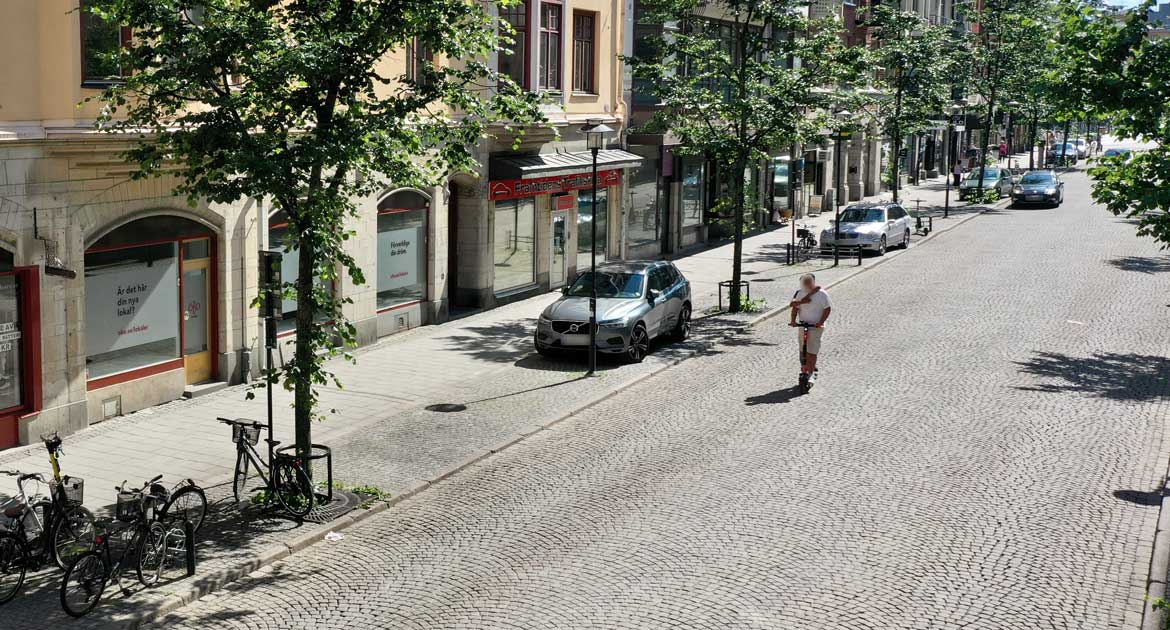 Storgatan 16 snett uppifrån med kullstensgata, bilar parkerade efter vägen och en person på elsparkcykel.