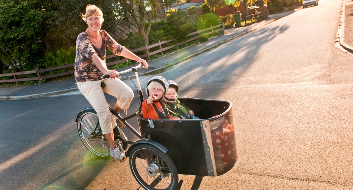 Kvinna som cyklar. Två små barn sitter i lådan framtill på cykeln.